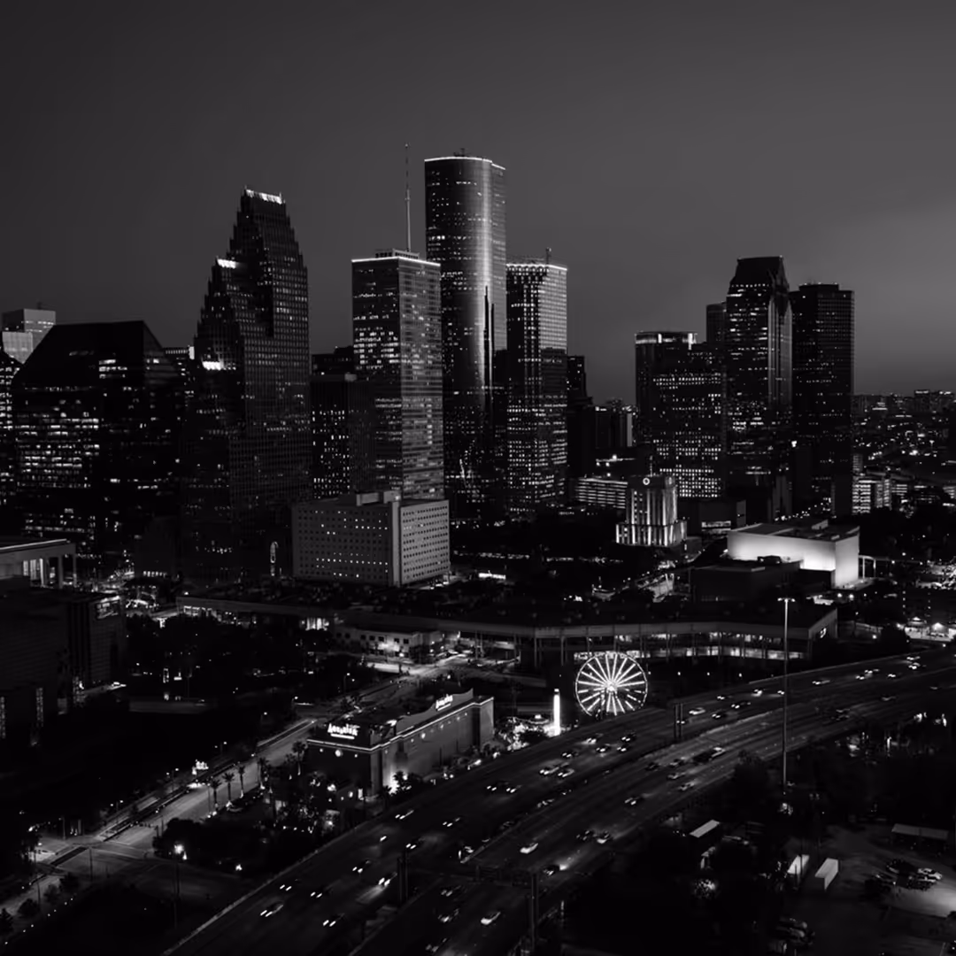 A black and white cityscape of Houston with tall modern skyscrapers glowing in the evening light and a highway in the foreground.