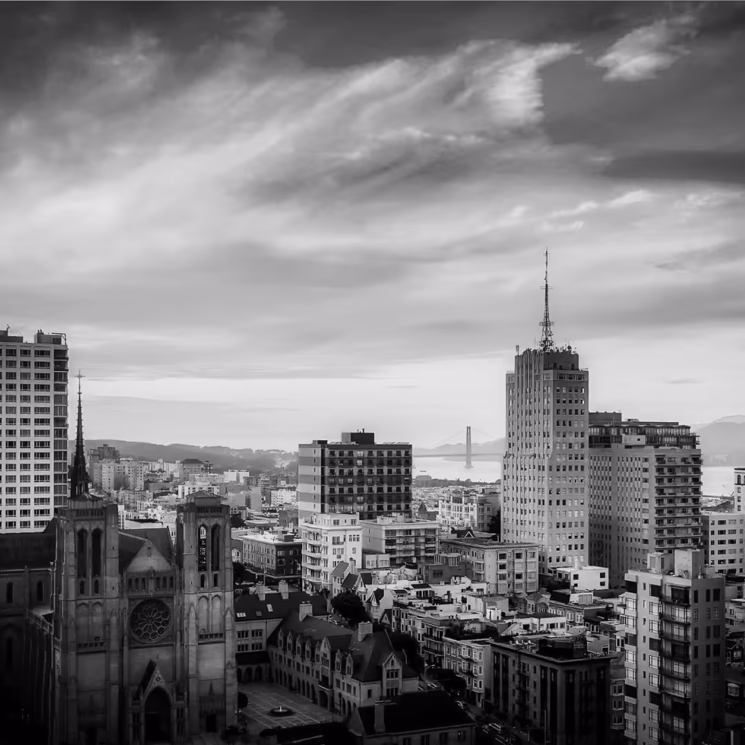A black and white view of San Francisco with Grace Cathedral in the foreground, tall buildings rising across the city, and the Golden Gate Bridge faintly visible in the background.