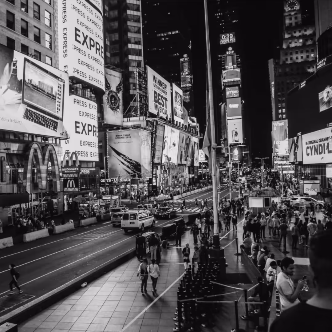 Black-and-white photo of Times Square filled with people, cars, and large illuminated billboards lining the busy streets.