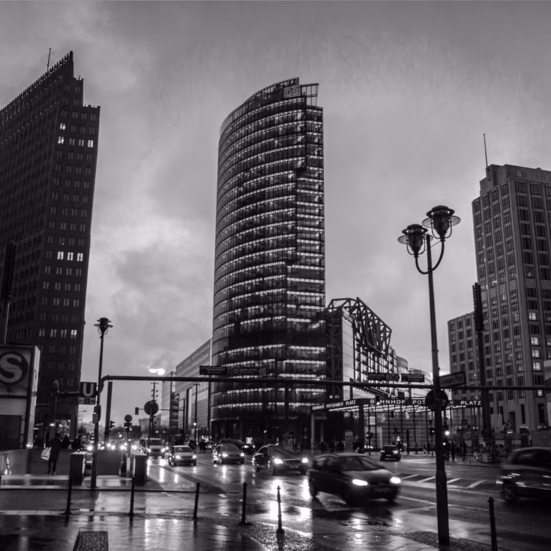 Black and white photo of a modern Berlin street scene with the curved DB Tower lit up, cars moving in the rain, and surrounding skyscrapers at dusk.