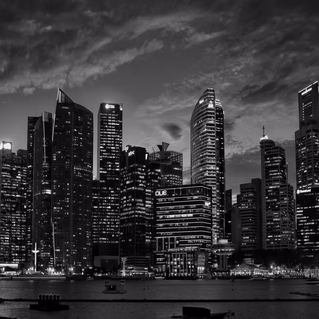 Black-and-white view of Singapore’s illuminated skyline at dusk, with modern skyscrapers including UBS and OUE Tower reflecting on the water.