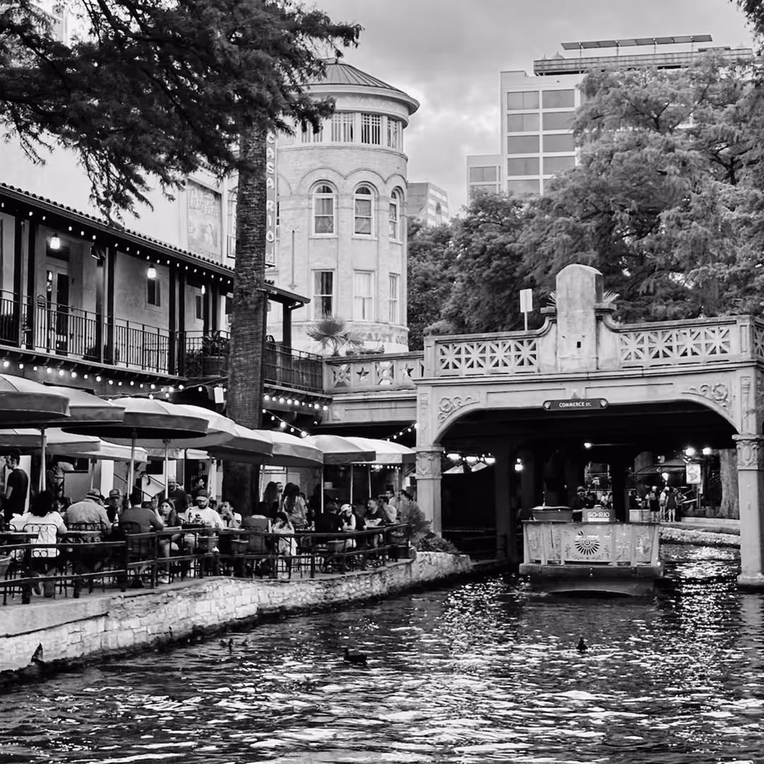 Black and white photo of San Antonio’s River Walk with outdoor dining under umbrellas, a tour boat passing under a decorative bridge, and historic buildings in the background.