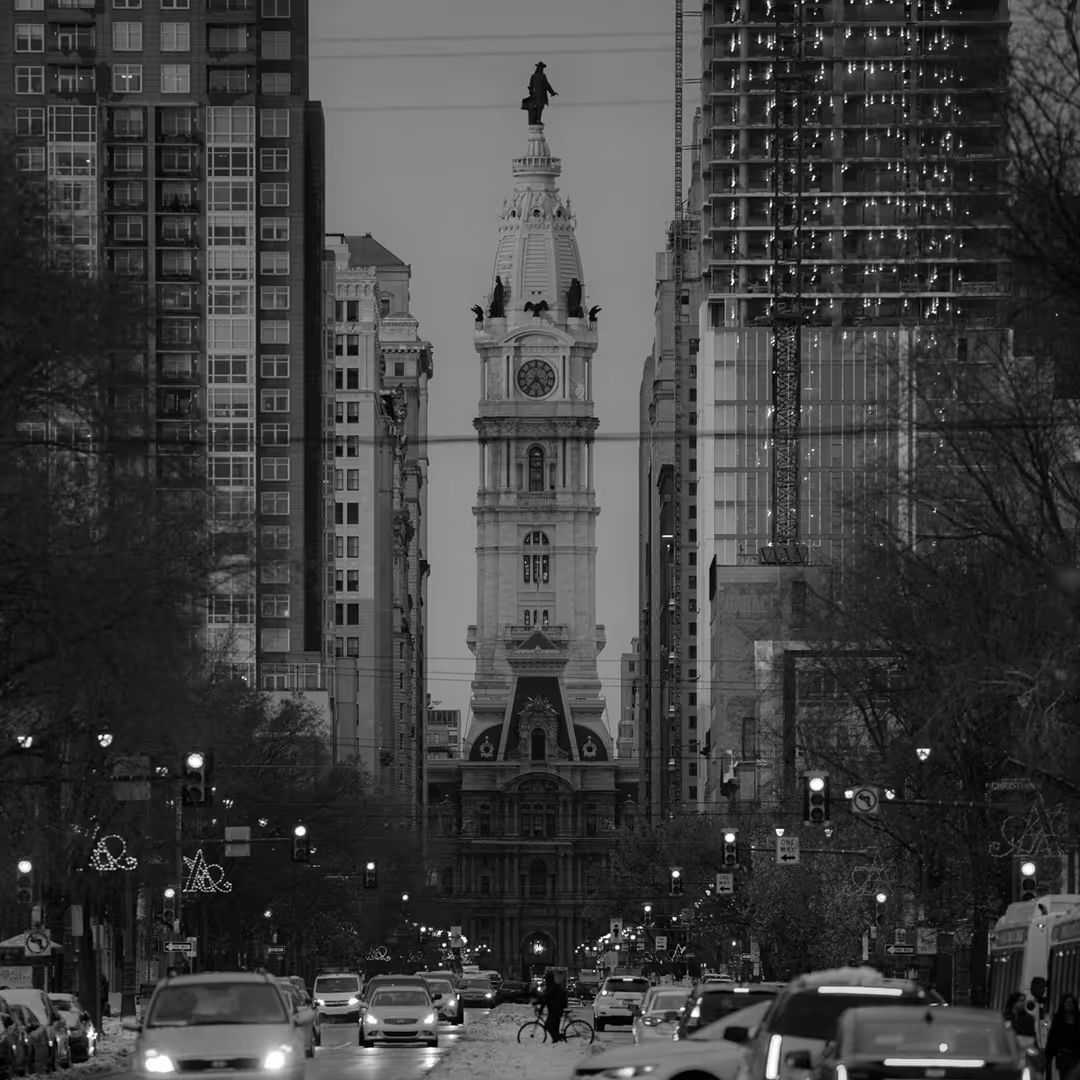 Street-level view in black-and-white of Philadelphia’s City Hall with William Penn’s statue on top, framed by tall buildings and traffic below.