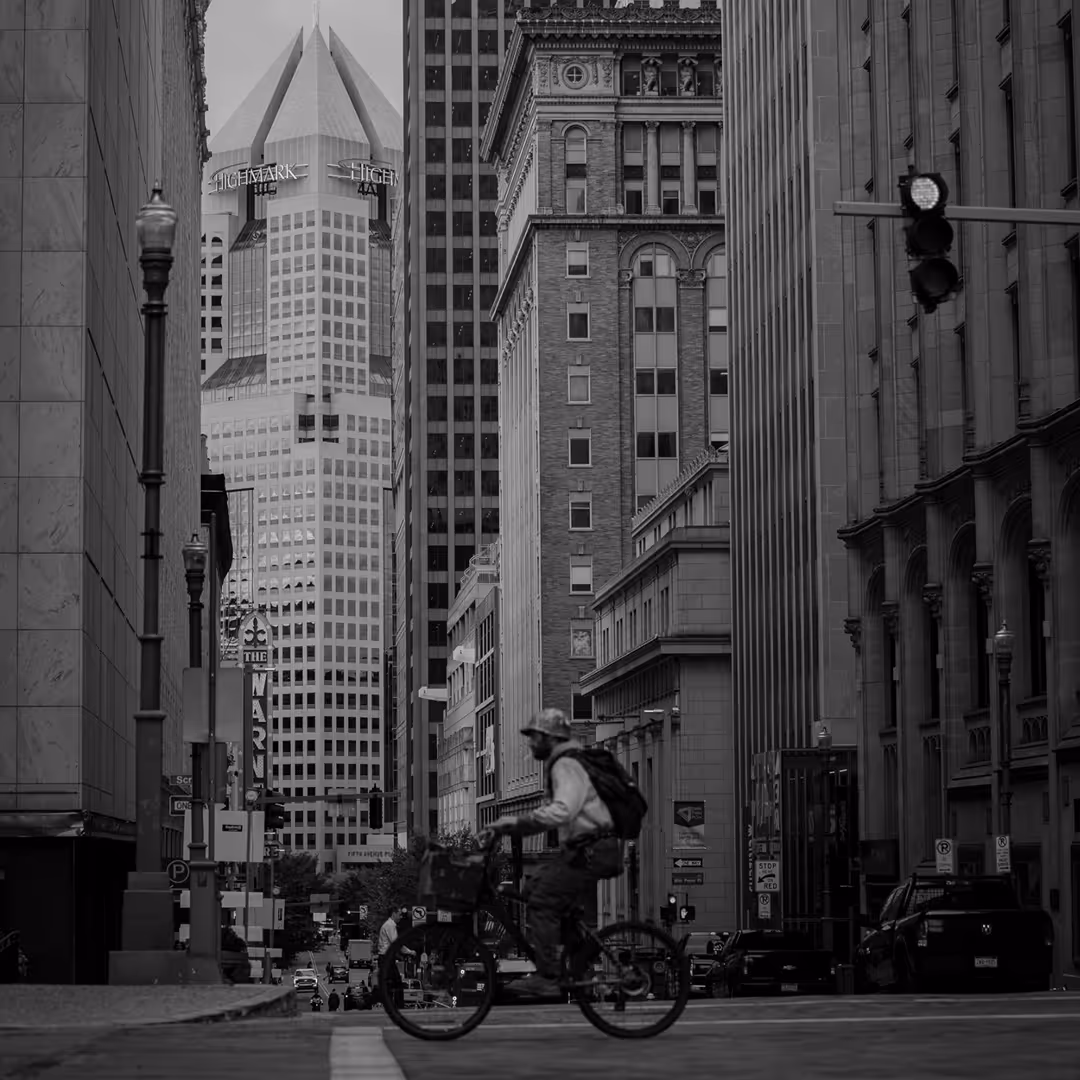 A black-and-white city street view with a bicyclist in the foreground, surrounded by tall buildings including the Highmark building with its pyramid-shaped top.