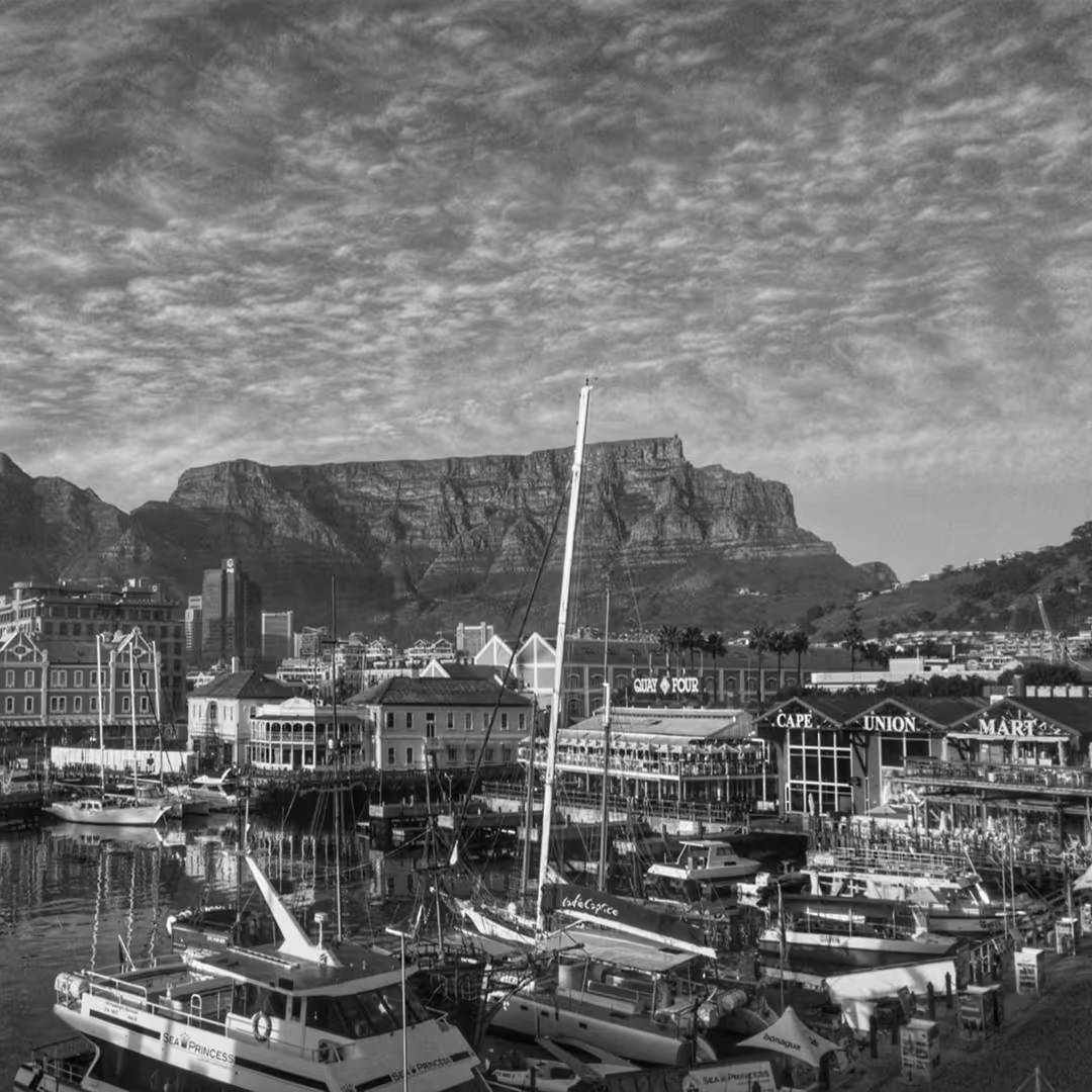 Black-and-white image of Cape Town’s V&A Waterfront, with boats docked in the harbor and Table Mountain rising in the background.