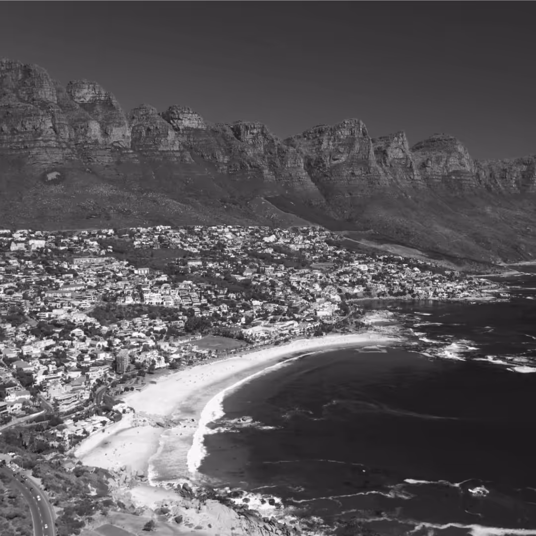 Black-and-white aerial view of Cape Town’s coastline with sandy beaches, residential areas, and the rugged peaks of the Twelve Apostles mountain range.