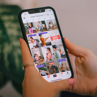 Close-up of a person browsing a TikTok-style video feed on a smartphone, showing a grid of diverse creators and short video clips with text overlays.