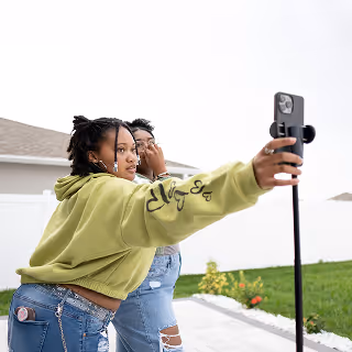 Two young women posing together for a selfie using a smartphone on a tripod, dressed casually in a backyard setting, capturing a moment of fun and authenticity.