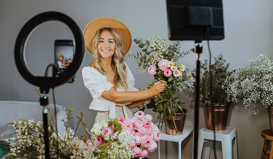 Woman smiling while arranging flowers on camera, using a smartphone and ring light setup for filming content in a cozy studio filled with floral arrangements.