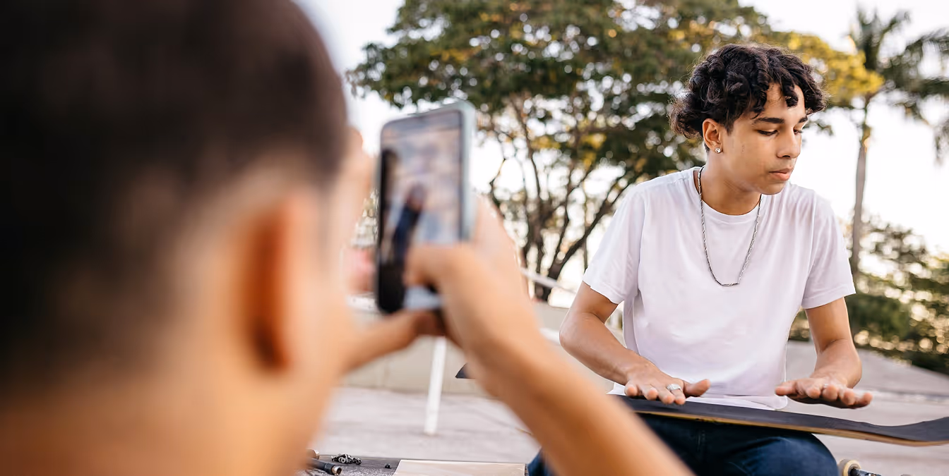 A teen plays the skateboard like a drum while a friend records the performance on a smartphone, capturing spontaneous creativity in a public park setting.
