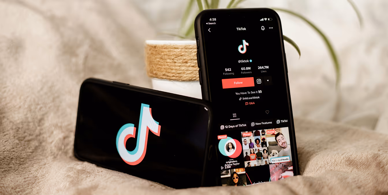 Two smartphones on a bed—one showing the TikTok logo and the other open to the official TikTok profile—framed beside a potted plant, illustrating dual-device social strategy.