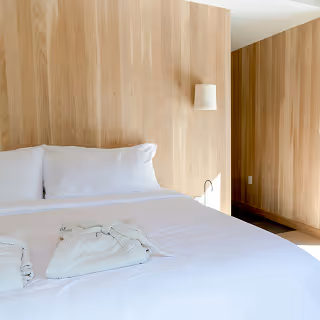 Bright and minimal bedroom with natural wood paneling, a neatly made white bed, and folded robes, conveying a clean, calming atmosphere.