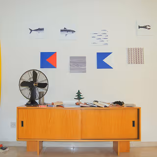 	Minimalist mid-century modern sideboard against a white wall, decorated with nautical and abstract artwork, books, and a vintage desk fan, emphasizing design details and personality.