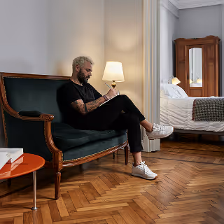 Man seated on a vintage green velvet loveseat in a cozy Airbnb suite, writing in a notebook with a bedroom visible in the background, illustrating thoughtful guest preparation.