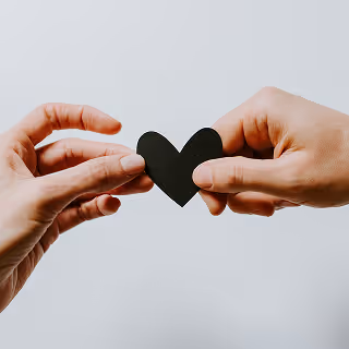 Two hands holding a small black heart shape together, symbolizing connection, care, or emotional bond.