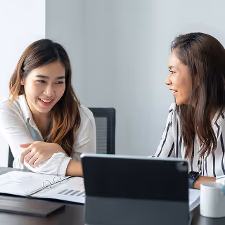 Two women in a modern office smiling and discussing business charts on a tablet, conveying friendly collaboration.