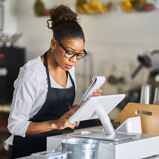 Female small business owner in apron reviewing a notebook while working at a point-of-sale system in a cozy shop.