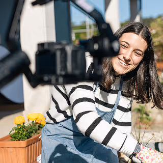 Smiling woman in striped sweater and apron kneeling by flower pots, being recorded with a camera on a gimbal outdoors.
