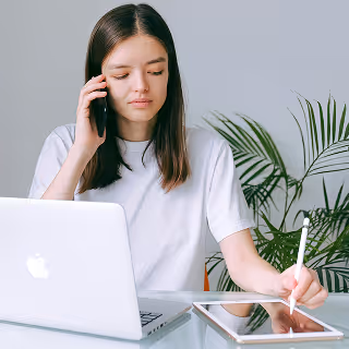 Young woman multitasking at a desk, speaking on the phone while writing on a tablet, with a laptop and plant nearby.