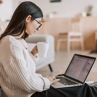 Woman sitting on the floor with a cup of coffee, working on a laptop in a cozy home setting.