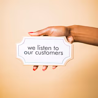 A hand holding a sign that reads “we listen to our customers” against a beige background.