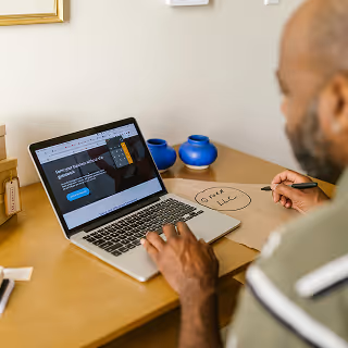 Entrepreneur using a laptop to browse a business service website while sketching out a brand name on paper, symbolizing brand planning.