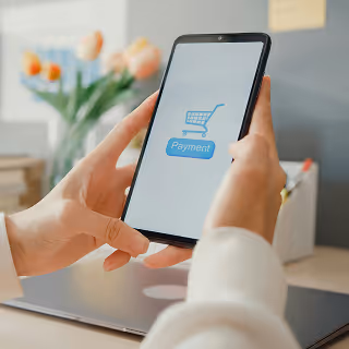 Person holding a smartphone with a “Payment” icon and a blue shopping cart symbol on the screen, indicating an online purchase process.