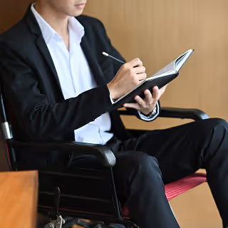 Businessperson in a wheelchair dressed in a black suit taking notes in a notebook, representing inclusivity and professional focus.