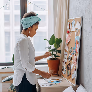 Creative young woman wearing a turquoise head wrap pins clippings to an inspiration board in a home studio with natural light and potted plants.