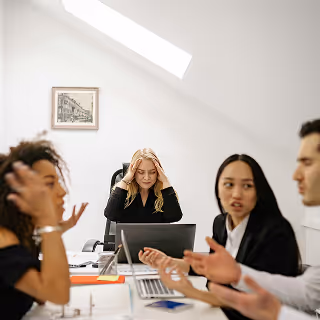 Four businesspeople engaged in a heated discussion around a table, while a blonde woman at the head appears stressed, holding her head in frustration.