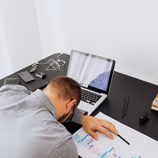 Frustrated businessman with head down on a desk covered in charts and graphs, suggesting burnout or overwhelming stress at work.