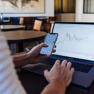 Person using both a smartphone and laptop to analyze cryptocurrency market charts in a café setting.