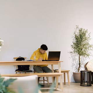 Man in a yellow hoodie working on a laptop and writing notes at a long wooden table in a bright, minimalist room with potted plants.