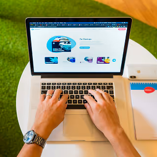 Person using a MacBook on a round white table, browsing a startup services platform, with a notepad, pen, and iPhone nearby.