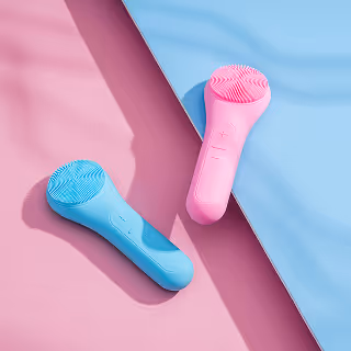 Flat lay of two silicone facial cleansing brushes in pastel pink and blue, placed diagonally across a pink and blue background.