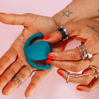 Hands adorned with rings and tattoos holding a teal, modern wellness device with curved extensions, against a pink background.