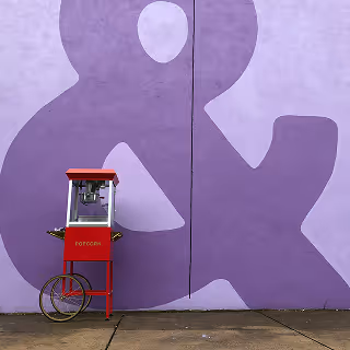 Red vintage popcorn cart standing on a sidewalk against a large lavender wall with a giant ampersand symbol painted on it.