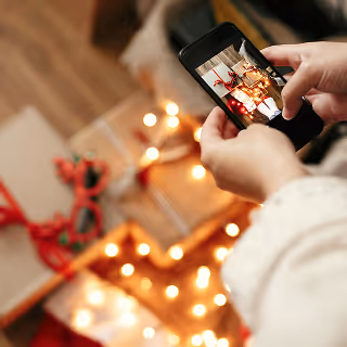 Close-up of a person taking a photo of wrapped Christmas presents and holiday lights using a smartphone.
