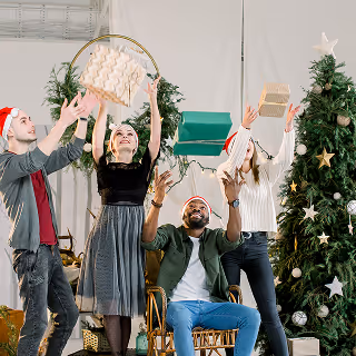 Group of four people in festive Santa hats tossing wrapped gifts into the air in front of Christmas trees.