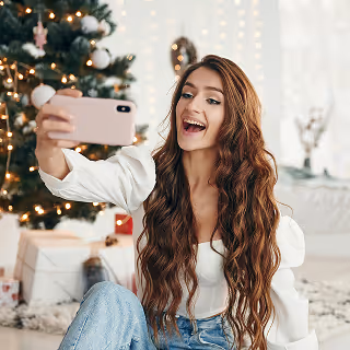 Smiling woman sitting in front of a decorated Christmas tree, holding a smartphone to take a holiday selfie.