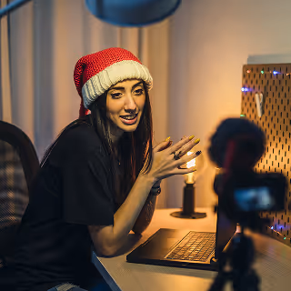 Young woman in a Santa hat vlogging in front of a laptop, speaking animatedly to a camera in a warmly lit holiday-themed room.