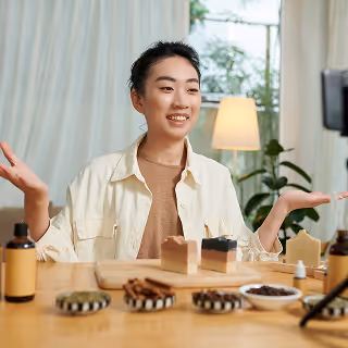 Smiling woman sitting at a table livestreaming handmade soap products and natural ingredients to a camera in a well-lit, cozy room.