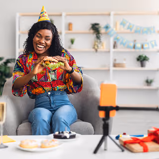 Cheerful woman in a party hat celebrating her birthday while creating social media content with a smartphone tripod, surrounded by cake, drinks, and festive decorations.