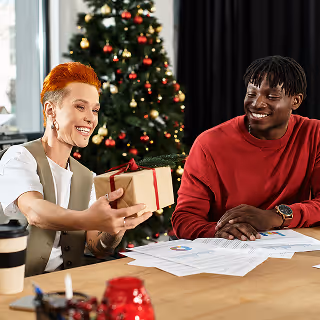 Two coworkers in a festive office setting exchanging a gift beside a decorated Christmas tree, with paperwork on the table.