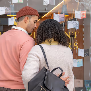 Couple standing close together, peering through a store window decorated with product cards and warm lighting, suggesting holiday or boutique shopping.