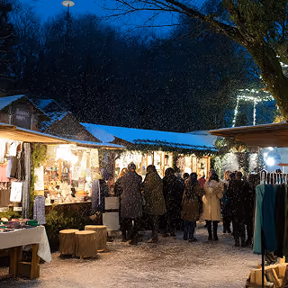 Crowds of people shop at a cozy outdoor holiday market in the evening, with string lights illuminating wooden stalls and snow falling softly.