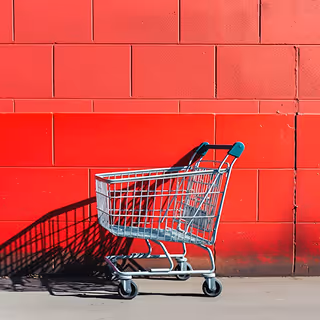 Empty metal shopping cart with teal handles parked against a bold red brick wall, casting a clear shadow on the pavement.