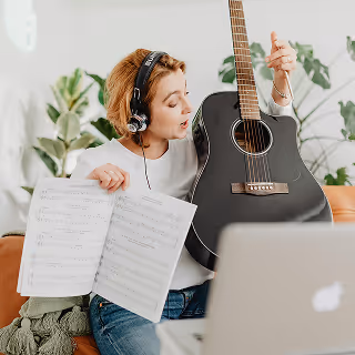 A woman wearing headphones holds a black acoustic guitar and sheet music while watching an online lesson on a laptop, seated on a couch in a cozy indoor space.