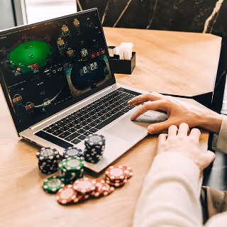Close-up of a person’s hands on a laptop showing an online poker game, with poker chips scattered on the table, conveying interactive online gambling.