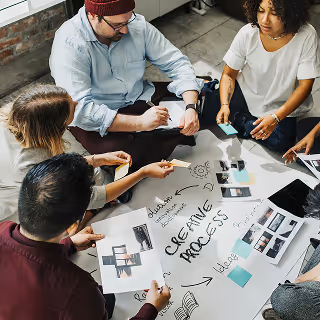 Team of creatives sitting on the floor collaborating on a project plan – A group of diverse individuals working together on a creative process diagram, surrounded by photos and sticky notes.
